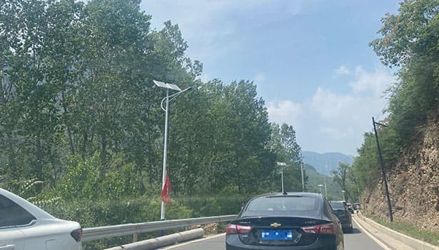 Solar LED street lights installed along a mountain road surrounded by lush green trees and passing cars
