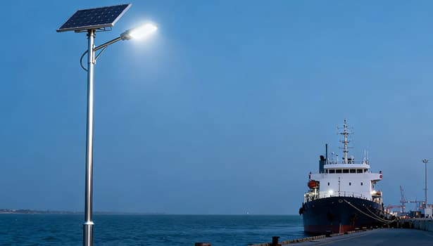 Solar street light illuminating a port dock area at dusk, with a cargo ship moored nearby and the sea in the background.