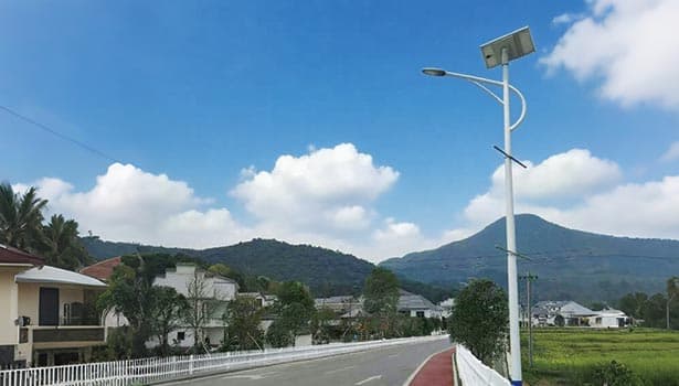 Solar street light installed along a residential area road, with houses, greenery, and mountains in the background under a blue sky